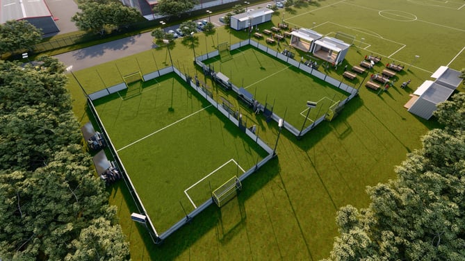 Aerial view of two adjacent 5-a-side soccer courts with players, seating areas, and nearby picnic tables, surrounded by trees and larger soccer fields.