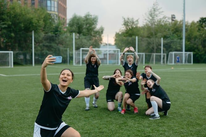 Smiling female soccer player taking a selfie with teammates posing and cheering in the background on a green soccer pitch.
