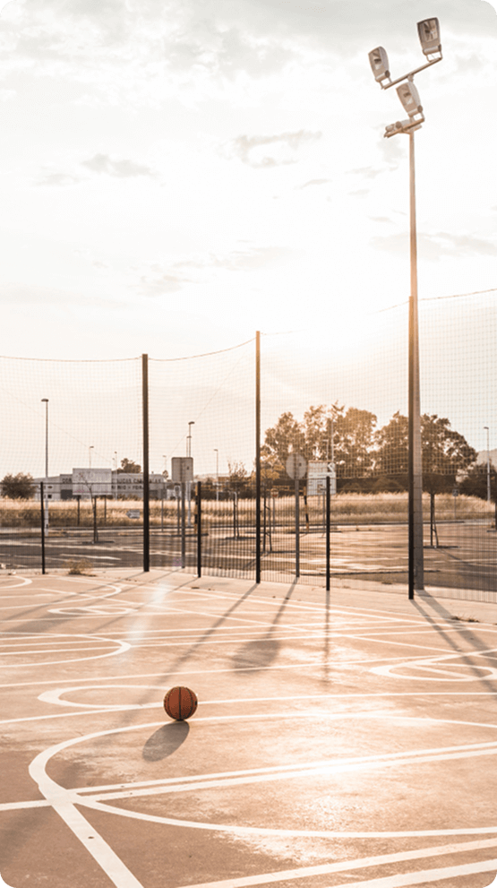 Sunset over an empty outdoor basketball court with a lone ball and tall lighting poles, evoking calm and community potential.