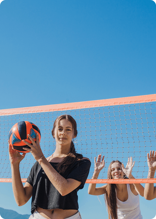 Group of young women smiling with a volleyball at an outdoor court net during sunset